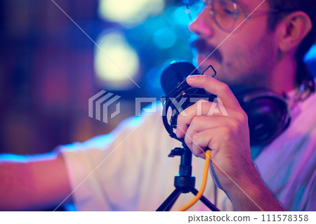 Side view portrait of young man in glasses, blogger talking to microphone while streaming online in neon light. Side view portrait of young man in glasses, blogger talking to microphone while streaming online in neon light. 111578358