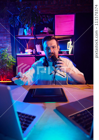 Annoyed young man, shouting to microphone while recording video-lesson for students in neon lit workspace at home. 111578374