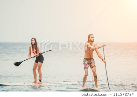 Sea woman and man on sup. Silhouette of happy young woman and man, surfing on SUP board, confident paddling through water surface. Idyllic sunset. Active lifestyle at sea or river. 111578784