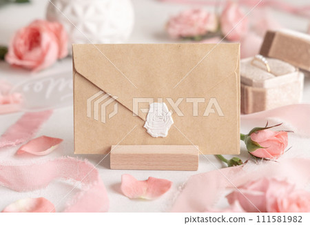 Blank sealed envelope near light pink roses, petals and silk ribbons close up, wedding mockup 111581982