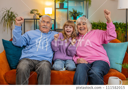Happy Caucasian grandparents child girl granddaughter watching TV celebrating success winning goal Happy Caucasian grandparents child girl granddaughter watching TV celebrating success winning goal 111582015