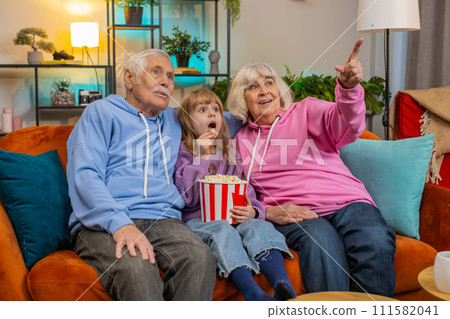 Child girl granddaughter grandfather grandmother eating popcorn watching movie film on home sofa 111582041