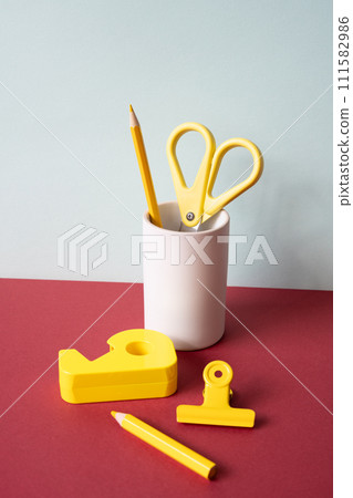 Office supply stationery. Yellow colored pencil, scissors, tape dispenser, clip on red desk. gray wall background. workspace Office supply stationery. Yellow colored pencil, scissors, tape dispenser, clip on red desk. gray wall background. workspace 111582986