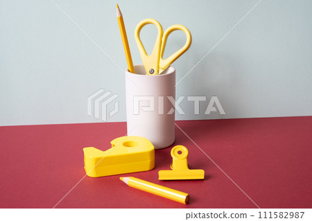 Office supply stationery. Yellow colored pencil, scissors, tape dispenser, clip on red desk. gray wall background. workspace 111582987