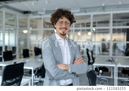 Portrait of a young mature male professional with curly hair confidently standing in a well-lit corporate office. 111583255