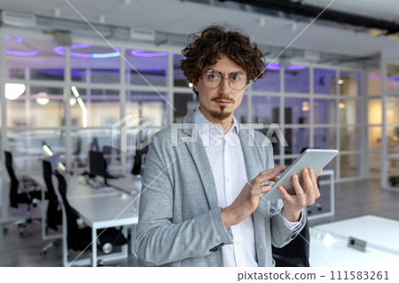 A young mature businessman with curly hair focuses on his tablet while working in a contemporary office setup, embodying professionalism. 111583261