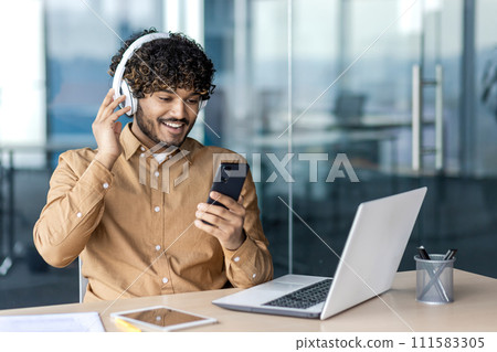 Smiling young Indian man sitting at desk in office with laptop and tablet, wearing white headphones, using mobile phone. 111583305