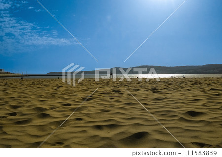 View of sand surface against blue sky background, with clouds, sun halo, reflection on ocean water. Unique natural areas, beaches, deserts. Place where all elements of Nature interact with each other 111583839