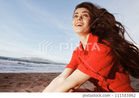 Serene Sunset: Beautiful Woman Sitting on the Beach, Enjoying Freedom and Vacation Bliss in the Warm Summer Evening 111583862