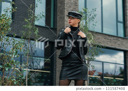 Cool trendy beauty young stylish woman model with fashion sunglasses in green dress with leather black jacket posing on the street near metal lines wall 111587408