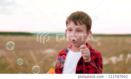 Boy spends day blowing soap bubbles in cultivated field. Boy child stands in nature savouring simple pleasure of soap bubbles. Boy in checkered shirt playing with soap bubbles stand in field Boy spends day blowing soap bubbles in cultivated field. Boy child stands in nature savouring simple pleasure of soap bubbles. Boy in checkered shirt playing with soap bubbles stand in field 111587602