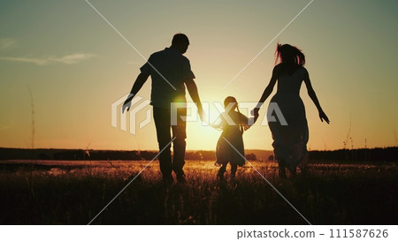 Small girl happily runs hand in hand with parents on field to release energy after day at kindergarten. Small girl loves to run across field at sunset having fun with papa and momma at sunset 111587626