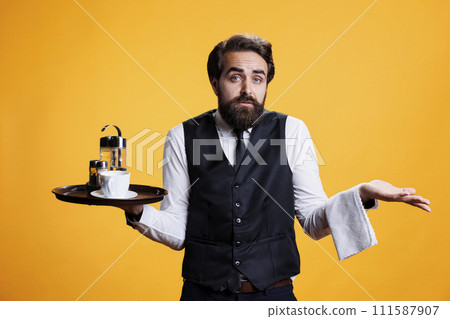 Doubtful employee with tray in hand raising shoulders and expressing uncertain feelings, yellow background in studio. Young man with waiter profession acting clueless and unsure on camera. 111587907