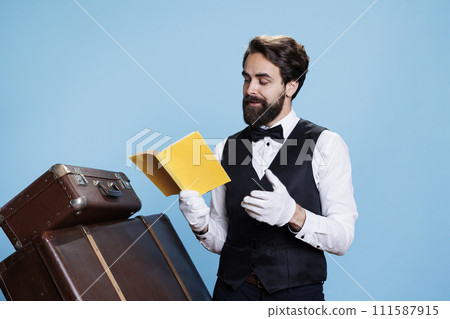 Stylish employee enjoys fiction book while on duty, taking care of trolley bags in studio. Male doorkeeper reading literature pages in front of camera, having fun with classic hobby. Stylish employee enjoys fiction book while on duty, taking care of trolley bags in studio. Male doorkeeper reading literature pages in front of camera, having fun with classic hobby. 111587915