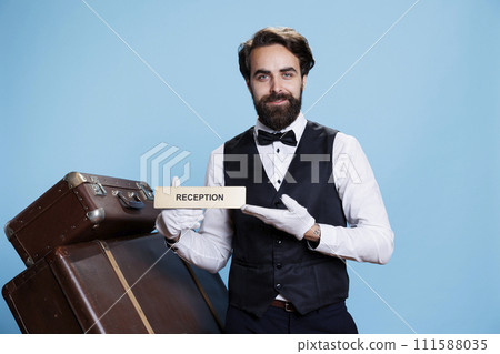 Hotel employee leading tourists in the right way using reception wall sign. Skilled professional bellboy posing on camera and holding front desk indicator, guiding guests and travellers. Hotel employee leading tourists in the right way using reception wall sign. Skilled professional bellboy posing on camera and holding front desk indicator, guiding guests and travellers. 111588035