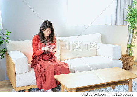 A woman in her 30s sits on a sofa in her room and operates a smartphone with a troubled expression on her face. A woman in her 30s sits on a sofa in her room and operates a smartphone with a troubled expression on her face. 111590138