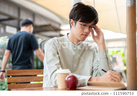 A focused Asian man is jotting down his ideas in a book while sitting at an outdoor table in a cafe. 111590733