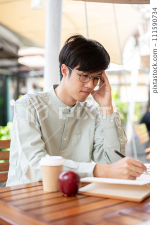 A focused Asian man is jotting down his ideas in a book while sitting at an outdoor table in a cafe. 111590734