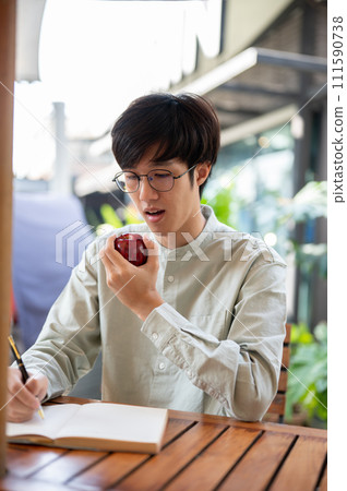 A handsome Asian man is eating an apple while writing down his ideas in a book at a cafe. A handsome Asian man is eating an apple while writing down his ideas in a book at a cafe. 111590738