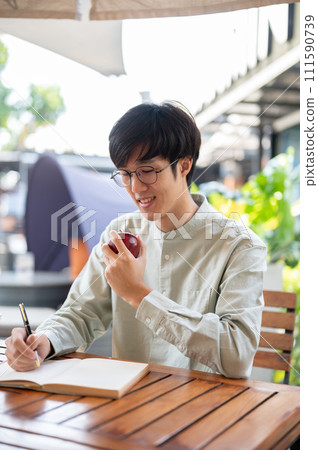 A handsome Asian man is eating an apple while writing down his ideas in a book at a cafe. 111590739