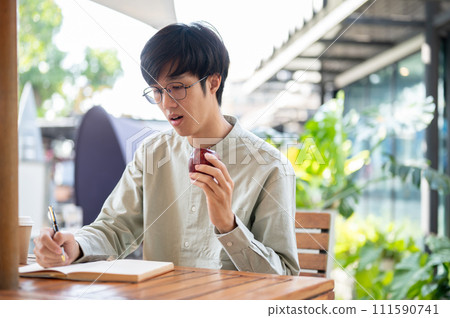 A handsome Asian man is eating an apple while writing down his ideas in a book at a cafe. A handsome Asian man is eating an apple while writing down his ideas in a book at a cafe. 111590741