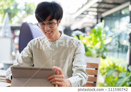 A happy young Asian man is working remotely at a coffee shop in the city, using his digital tablet. A happy young Asian man is working remotely at a coffee shop in the city, using his digital tablet. 111590742