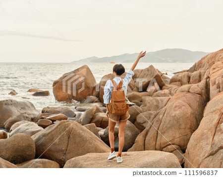 Tropical Sunset Adventure: A Happy Woman Walking on a Beach with a Backpack, Enjoying a Carefree Trip and Smiling at the Ocean Tropical Sunset Adventure: A Happy Woman Walking on a Beach with a Backpack, Enjoying a Carefree Trip and Smiling at the Ocean 111596837
