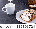 Milk is poured into a mug, next to a bun with poppy seed filling on a white plate, dark background 111598724