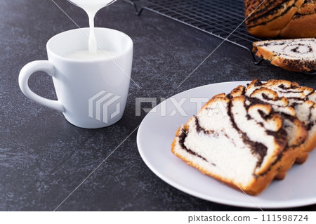 Milk is poured into a mug, next to a bun with poppy seed filling on a white plate, dark background Milk is poured into a mug, next to a bun with poppy seed filling on a white plate, dark background 111598724