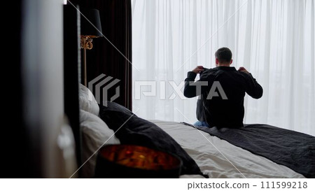 An adult man in a shirt in a room looks out the window. Businessman morning. A cozy room with a bed on which the guy sits with his back. A guy in a black shirt is sitting on the bed. An adult man in a shirt in a room looks out the window. Businessman morning. A cozy room with a bed on which the guy sits with his back. A guy in a black shirt is sitting on the bed. 111599218