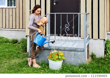Young woman with baby in her arms waters flower bed from garden watering can. 111599405