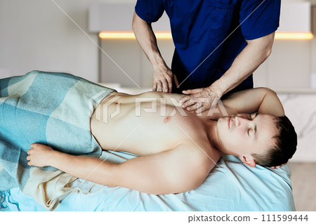 Mobile massage therapist kneads chest muscles of young man lying on massage table at home. Mobile massage therapist kneads chest muscles of young man lying on massage table at home. 111599444
