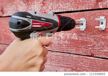 Using cordless screwdriver to install external pipeline holder latches on weathered red wood wall. Using cordless screwdriver to install external pipeline holder latches on weathered red wood wall. 111599605