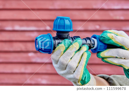 Tee PP screw joint fitting for drinking water clamp connector, plumber wearing protective gloves tightens clamping nut, on background of red wooden wall. Tee PP screw joint fitting for drinking water clamp connector, plumber wearing protective gloves tightens clamping nut, on background of red wooden wall. 111599678