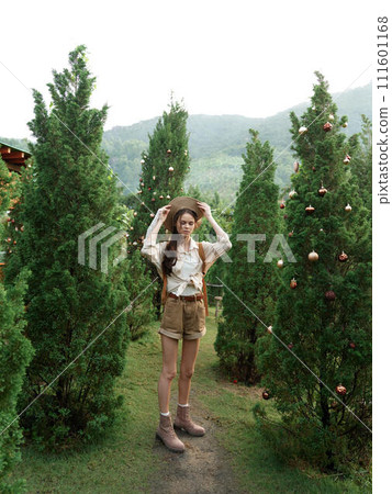 A woman wearing a hat and shorts standing in front of a large group of trees 111601168