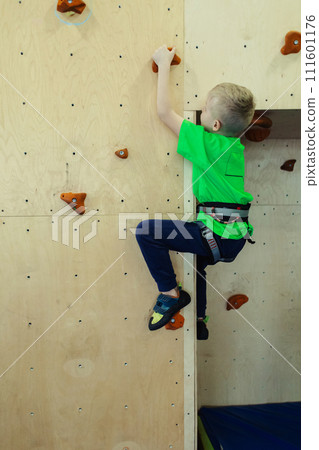 Rock climbing for children. Blond boy in a green shirt is climbing climbing wall. Bouldering class. Artificial track for safe activities and competitions. Mountaineering, safety system, special shoes 111601176