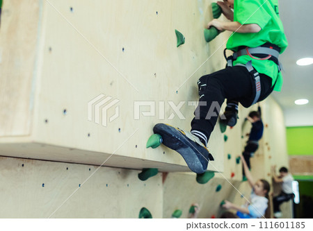 Rock climbing for children. Blond boy in a green shirt is climbing climbing wall. Bouldering class. Artificial track for safe activities and competitions. Mountaineering, safety system, special shoes 111601185