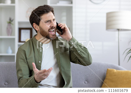 A bearded middle-aged man is engaged in an animated phone conversation, sitting alone on a sofa in a well-lit living room. A bearded middle-aged man is engaged in an animated phone conversation, sitting alone on a sofa in a well-lit living room. 111601679