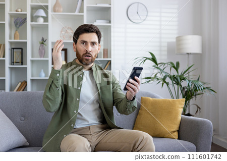 Adult man expressing shock and confusion while looking at his smartphone, seated comfortably on a living room sofa. 111601742