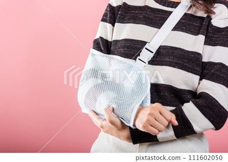 Confident businesswoman sporting a broken arm smiles warmly with an arm splint, receiving sling support from an excited Asian woman in a studio shot on pink background. 111602050