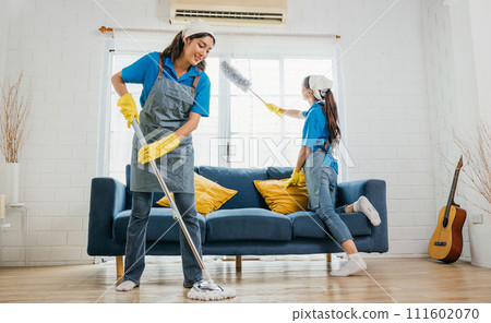 Two smiling women in uniform in messy living room working a team for house cleaning service. Using professional equipment they care for furniture sofa and hygiene. Housework is their routine. 111602070