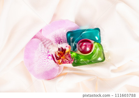 Gel liquid laundry detergent capsule with floral scent with orchid flower on fabric. Gel liquid laundry detergent capsule with floral scent with orchid flower on fabric. 111603788