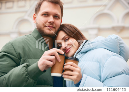 Lovely couple, young man and woman hugs and drinking coffee together while walking outdoor. First date. Lovely couple, young man and woman hugs and drinking coffee together while walking outdoor. First date. 111604453