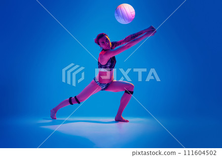 Concentrated athlete woman, beach volleyball player passes ball from below against gradient blue background in pink neon light. 111604502