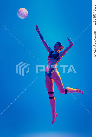 Athlete woman leaping to strike volleyball ball in sports attire in motion in mid-air against gradient blue background in pink neon light. 111604513