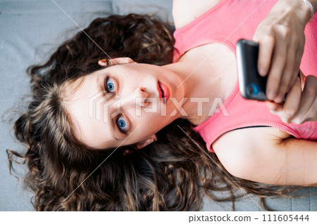Young woman with long curly hair lies on the sofa and scrolls social networks on her phone in a pink top, top view Young woman with long curly hair lies on the sofa and scrolls social networks on her phone in a pink top, top view 111605444