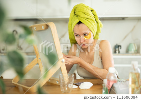 Middle-aged woman applying eye patch mask on her face sitting in front of the mirror Middle-aged woman applying eye patch mask on her face sitting in front of the mirror 111606094