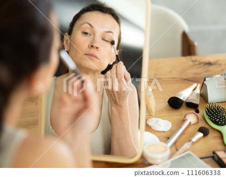Middle-aged woman using brush during eye makeup sitting in front of the mirror Middle-aged woman using brush during eye makeup sitting in front of the mirror 111606338