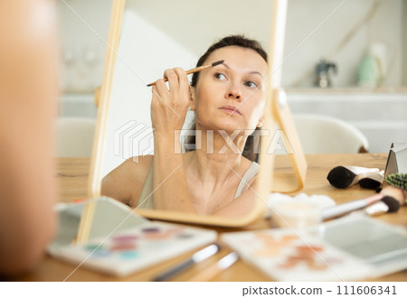 Middle-aged woman doing eyebrow make-up looking in the mirror Middle-aged woman doing eyebrow make-up looking in the mirror 111606341