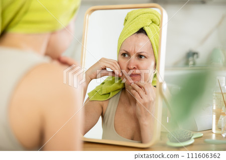 Dissatisfied woman with towel-wrapped hair scrutinizing facial skin in mirror 111606362